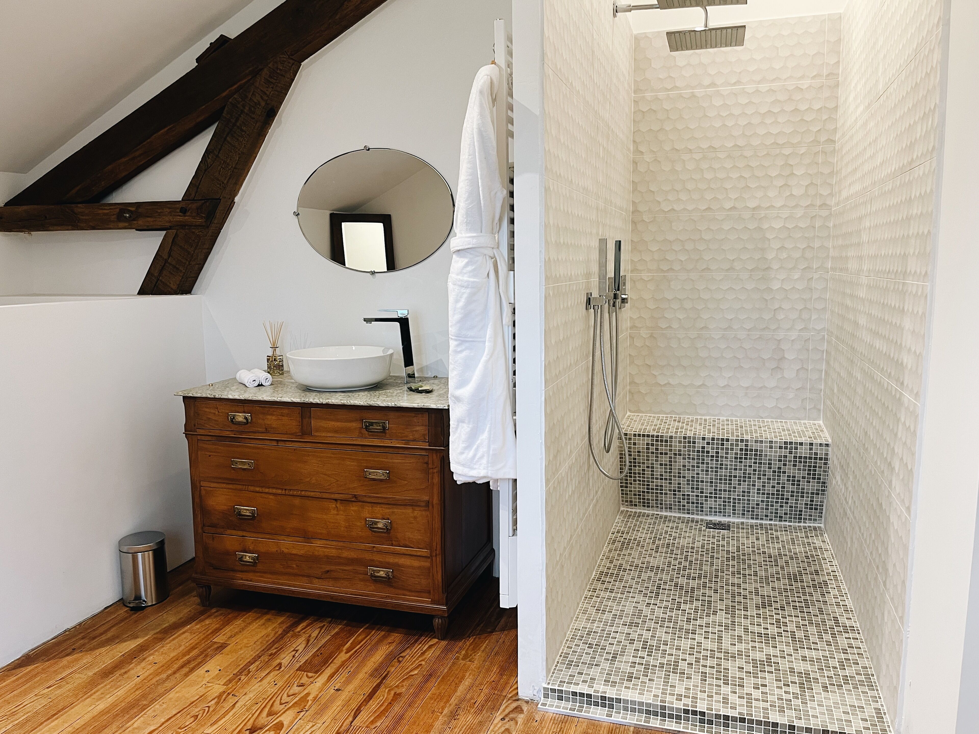 Un baño con tocador de madera, un espejo redondo, un lavabo blanco y una ducha con mosaicos.