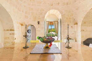 A dining table with a fruit bowl centerpiece, two candle holders, and a view of a garden through an archway. - Trulli Almaviva by Perle di Puglia (Cisternino)