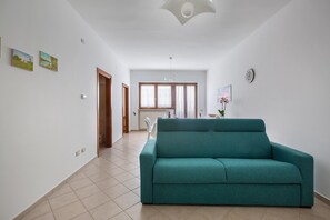 A spacious living room with a teal sofa, a dining area with a table and chairs, and a kitchen in the background.