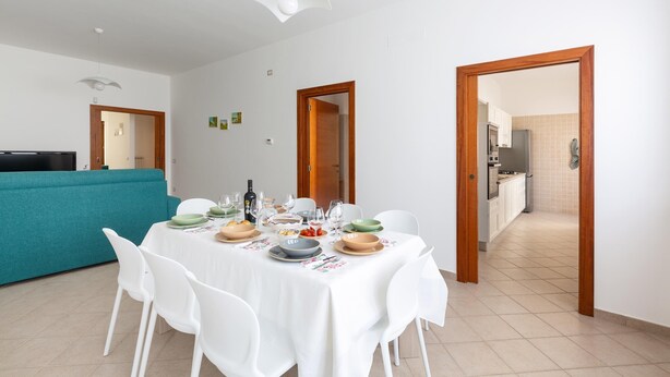 Un coin repas avec une table blanche dressée pour quatre personnes, entourée de chaises blanches. On aperçoit un canapé turquoise à l’arrière-plan, et une cuisine est visible à travers une porte ouverte.