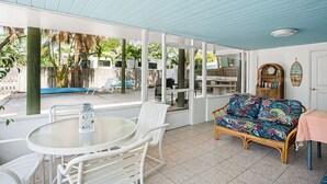 Een overdekt terras met een ronde tafel en stoelen, een rieten bank en een boekenkast.