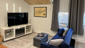 A modern living room with a flat-screen TV, a white entertainment unit, a blue sofa, and a large window with grey curtains.