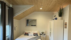 A bedroom with a sloped ceiling, a bed with white bedding and decorative pillows, a nightstand with a clock, a wardrobe, and a window with curtains.