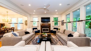 A spacious living room with two sofas, a coffee table, and a fireplace. There is a television mounted above the fireplace and large windows on the right side. - 2027 OPEN! Union Pier Home With Pool and Hot Tub! Close To Private Beach! (Union Pier)