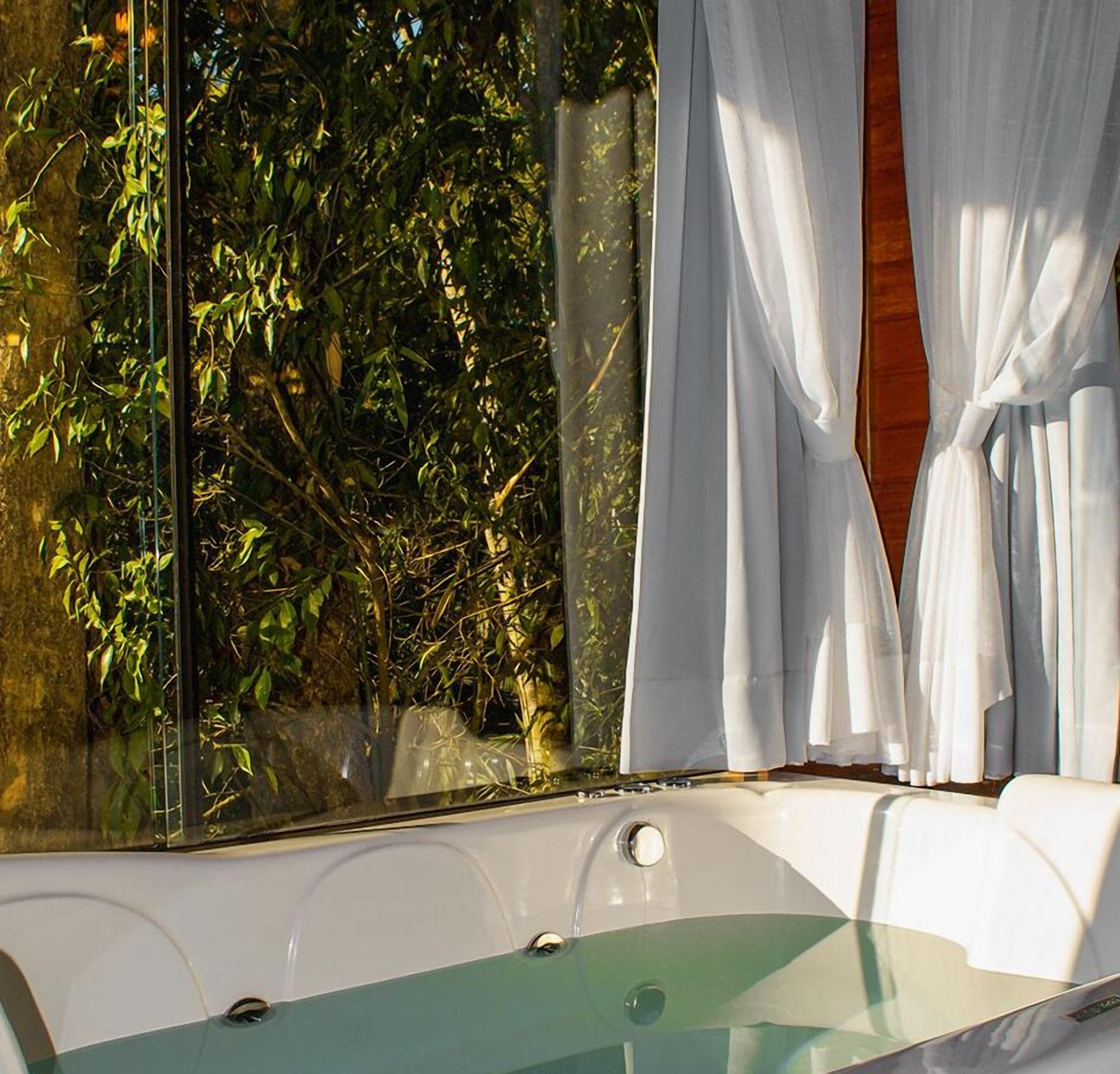 A modern bathroom with a glass-enclosed tub, white curtains, and a view of lush greenery outside.