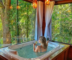 A woman in a hot tub with a glass of wine, surrounded by lush greenery and curtains.