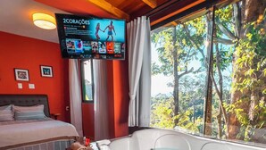 Habitación de hotel con cama, un ventanal con vistas a árboles, un televisor montado en la pared y una bañera de hidromasaje.