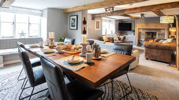 A spacious living area with a wooden dining table set for a meal, a fireplace, and a cozy seating area with a sofa and chairs. - Longlands Farm Cottage (Grange-over-Sands)