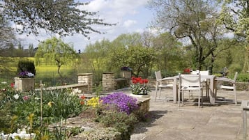 A garden patio with stone flooring, a table and chairs, and various flowering plants.