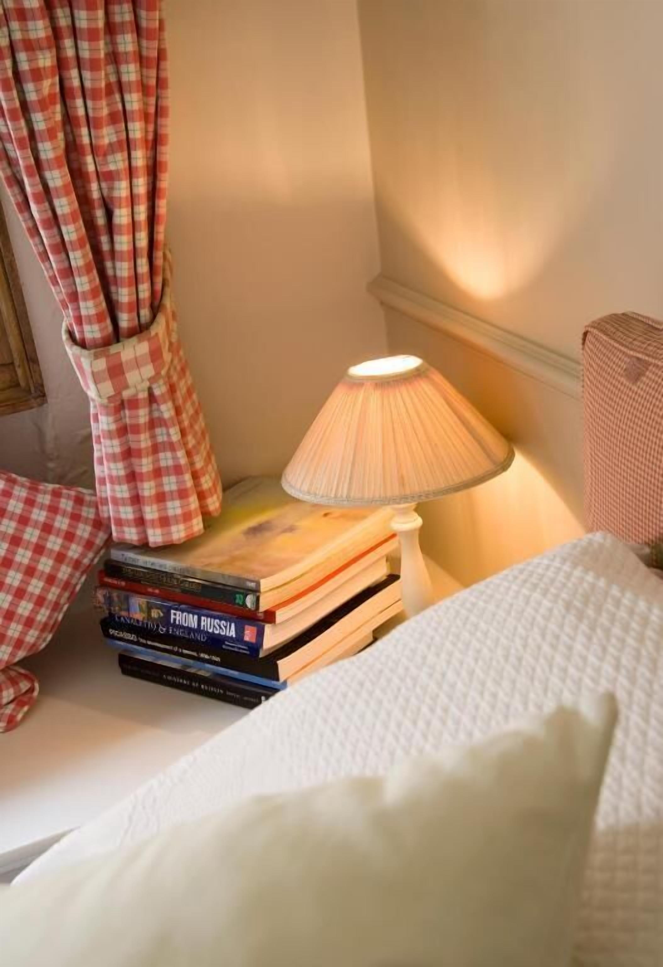En pænt redt seng med et hvidt dynebetræk, en stak bøger, heriblandt 'From Russia', en lampe med en beige skærm og en ternet gardin.