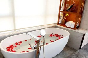 Een bad vol water en rozenblaadjes, een houten plank met handdoeken en toiletartikelen, en een marmeren vloer.