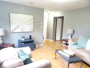 A living room with a large abstract painting, a blue entertainment unit, a wooden coffee table, and a beige sofa with a teal throw pillow. - Suzie's Place (Cornwall)