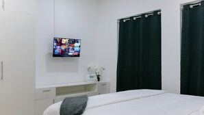 Een slaapkamer met een bed, een aan de muur bevestigde televisie, een klein tafeltje met een bloevas en donkere gordijnen.