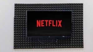 Een televisie bevestigd aan een muur, met een zwarte, structuurrijke omlijsting en het woord 'NETFLIX' in rode letters. Daaronder een witte ronde tafel met een zwarte lantaarn en een vaas met bloemen.