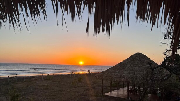 A beach view with a sunset, a thatched-roof structure, and a fenced area.