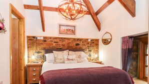 Un dormitorio con pared de piedra, techo de madera, cama con manta púrpura, mesita de noche con lámpara y ventana con cortinas.