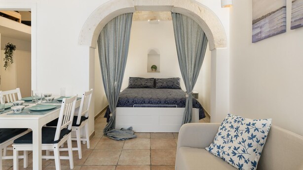 A room with a bed under an arched window, a dining table with chairs, and a sofa with a patterned pillow. - Cozy Historic Home by Wonderful Italy (Ostuni)