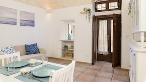 A dining area with a table set for two, a sofa with patterned cushions, and a kitchen area with a white cabinet and a window. - Cozy Historic Home by Wonderful Italy (Ostuni)