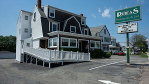 Un bâtiment à deux étages, à la façade blanche, doté d’un porche. On peut voir une enseigne indiquant le restaurant et l’auberge RHEAS, qui propose une offre spéciale pour le petit-déjeuner.