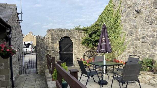 Eine Terrasse im Freien mit Steinmauern, einem schwarzen Tor, einem Tisch mit Stühlen und einem Sonnenschirm.