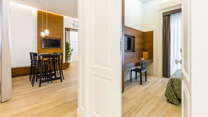 Una camera d'albergo moderna con pavimento in legno, un'area pranzo con tavolo e sedie, una TV a parete e un letto con copripiumino a motivi verdi.