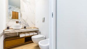Un bagno moderno con parete in marmo, lavandino bianco, mobile vanitario in legno, un water e un bidet.