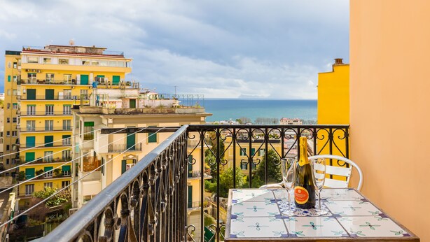 A balcony with a table, a bottle of wine, and a glass. The balcony overlooks a cityscape with buildings and a view of the sea. - B&B Civico 33 - Parthenope Qdpl (Napoli)