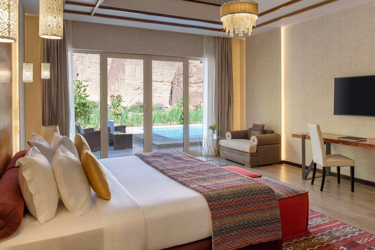 A hotel room with a bed, a desk, a TV, and a view of a pool and outdoor area.