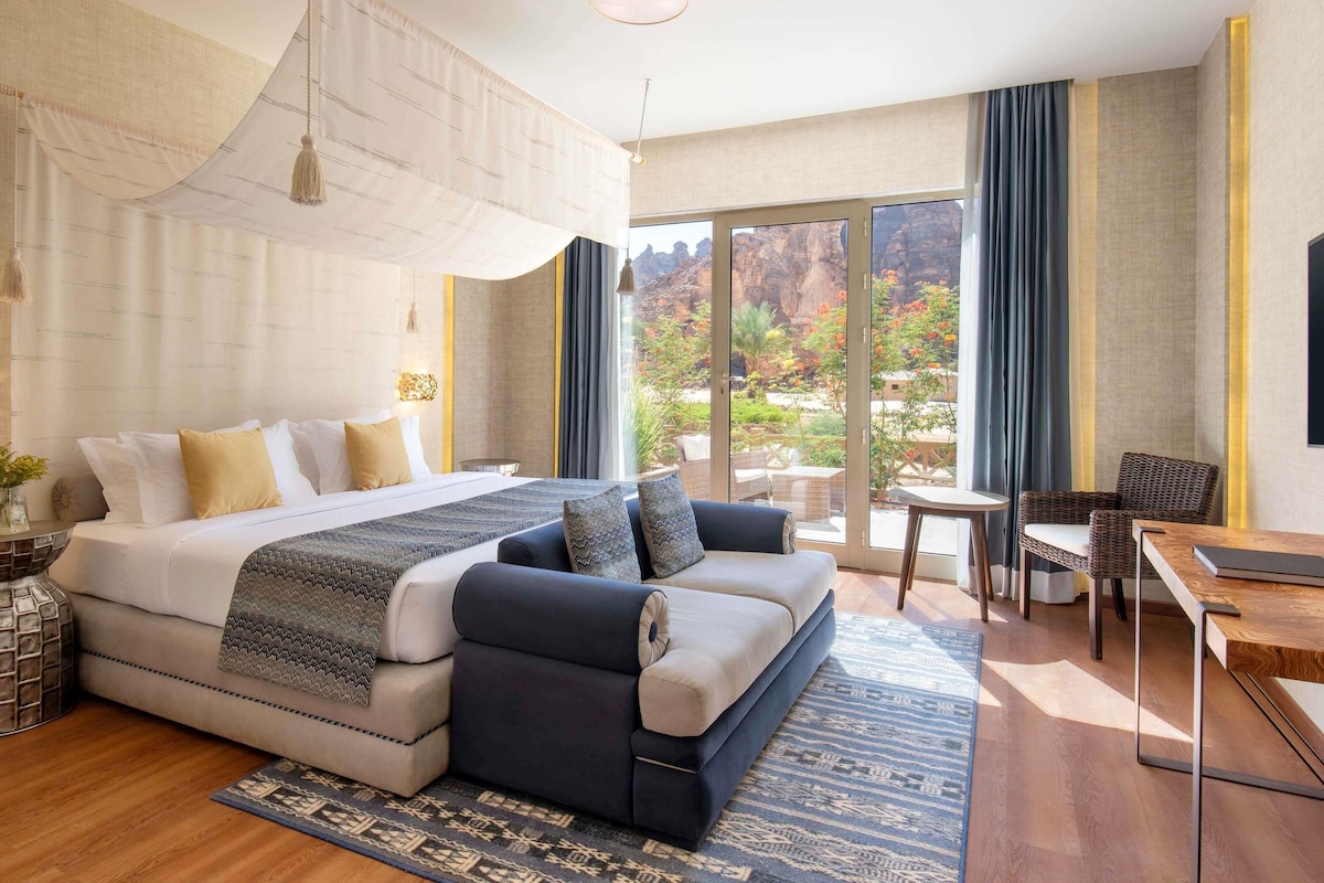 A modern hotel room with a large bed, a sofa, a dining table, and a balcony with a view of the outdoors.