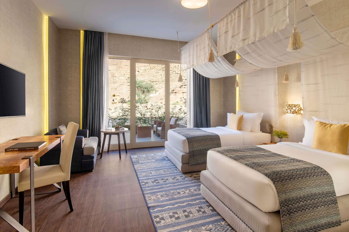 A modern hotel room with a desk, two beds, a large window, and a view of the outdoors.
