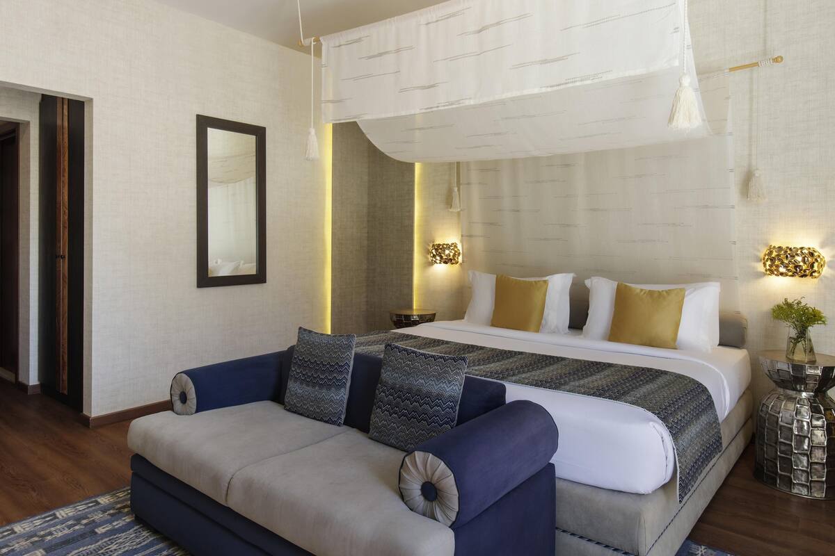 A modern bedroom with a large bed, a sofa, a mirror, and wall-mounted lights.