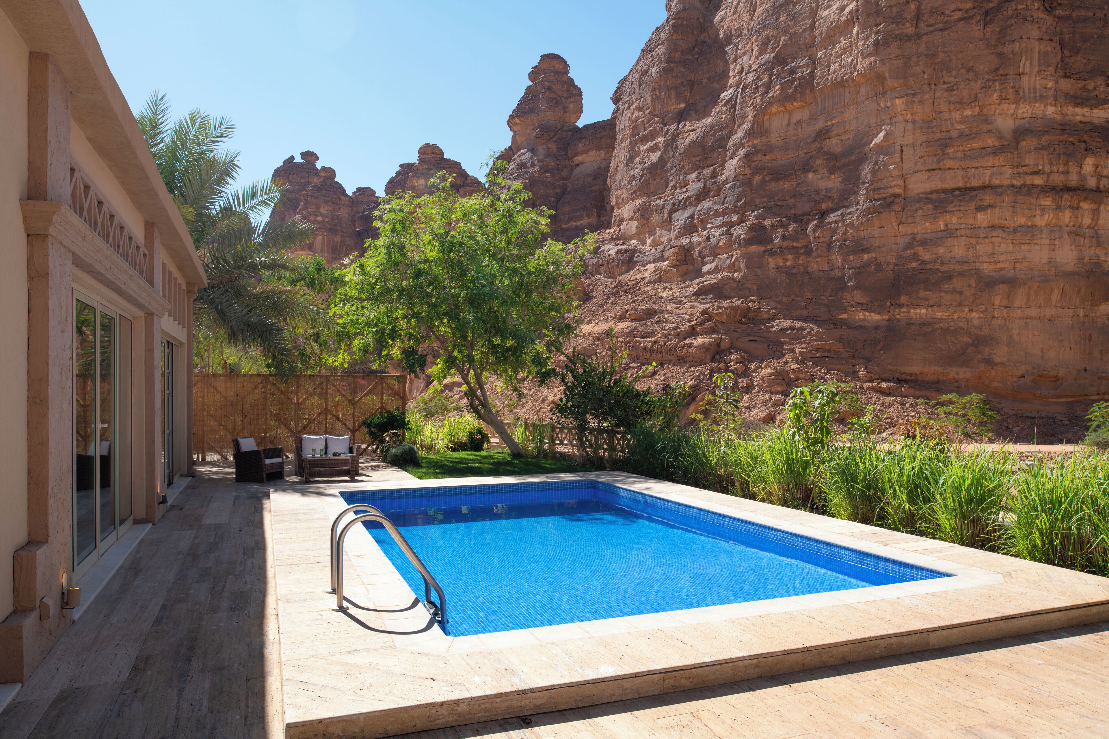 A rectangular swimming pool with a clear blue water, surrounded by a wooden deck and a stone wall.