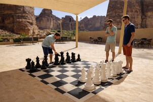 Un juego de ajedrez al aire libre sobre un suelo de cuadros, con sillas y mesas al fondo.