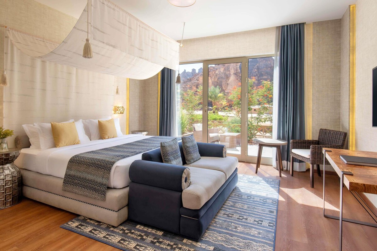 A modern hotel room with a large bed, a sofa, a dining table, and a balcony with a view of the outdoors.