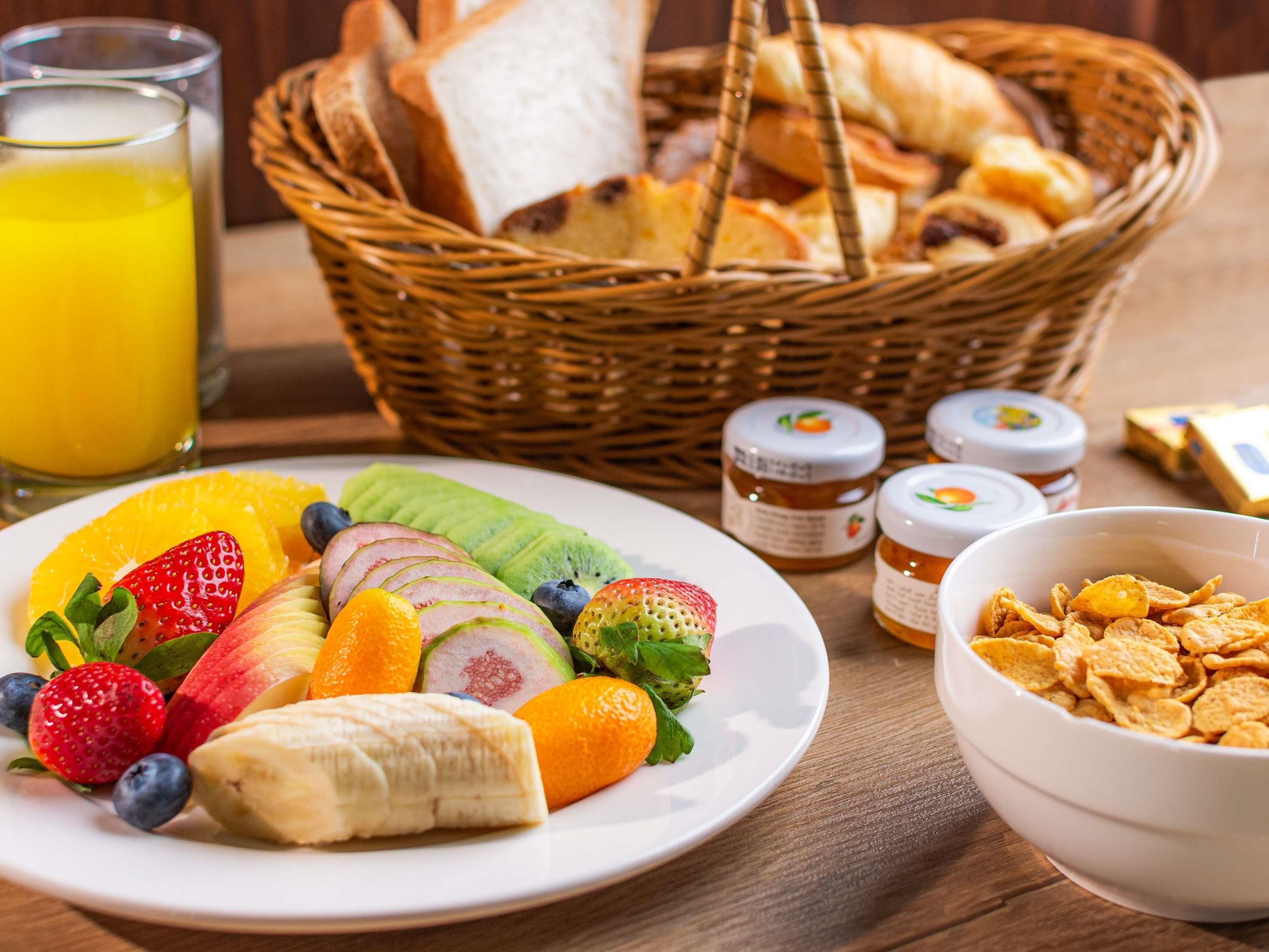 A breakfast setting with a plate of fruit, a bowl of cereal, a basket of bread, and a glass of orange juice.