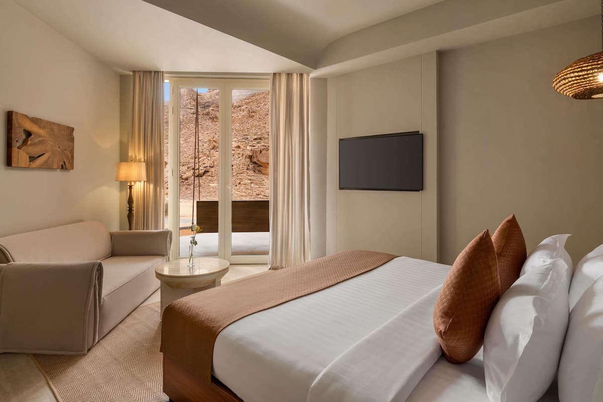 A hotel room with a bed, a sofa, a small table with a lamp, a TV, and a view of a rocky landscape through a window.