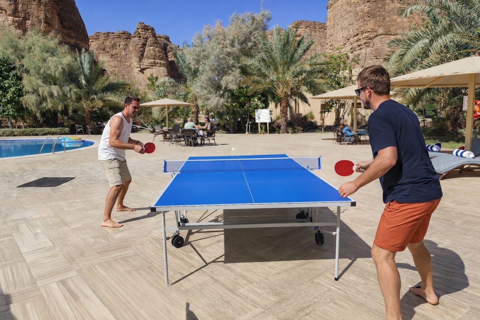 Two people playing table tennis on a poolside with a pool and outdoor seating area.
