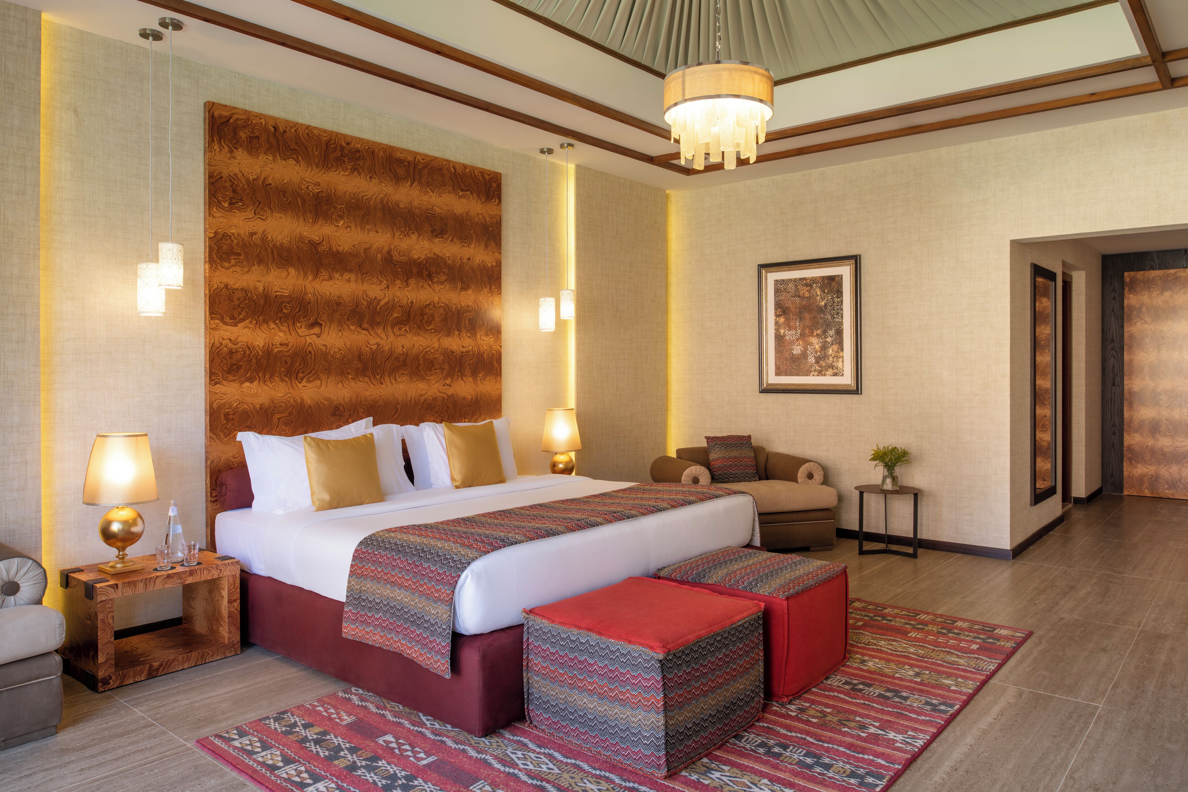 A hotel room with a large bed, bedside tables, a sofa, a patterned rug, and a chandelier.