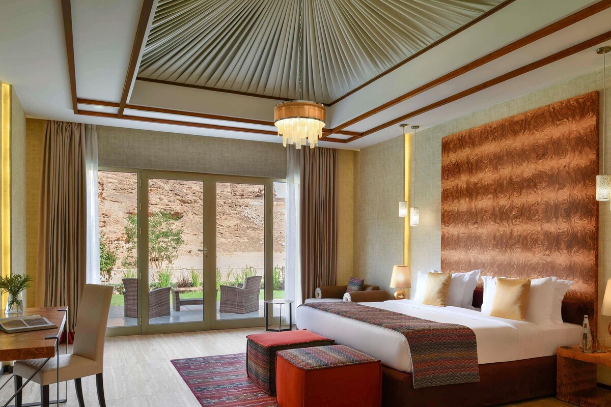 A hotel room with a large bed, a desk with a chair, a dining table, and a view of the outdoors.