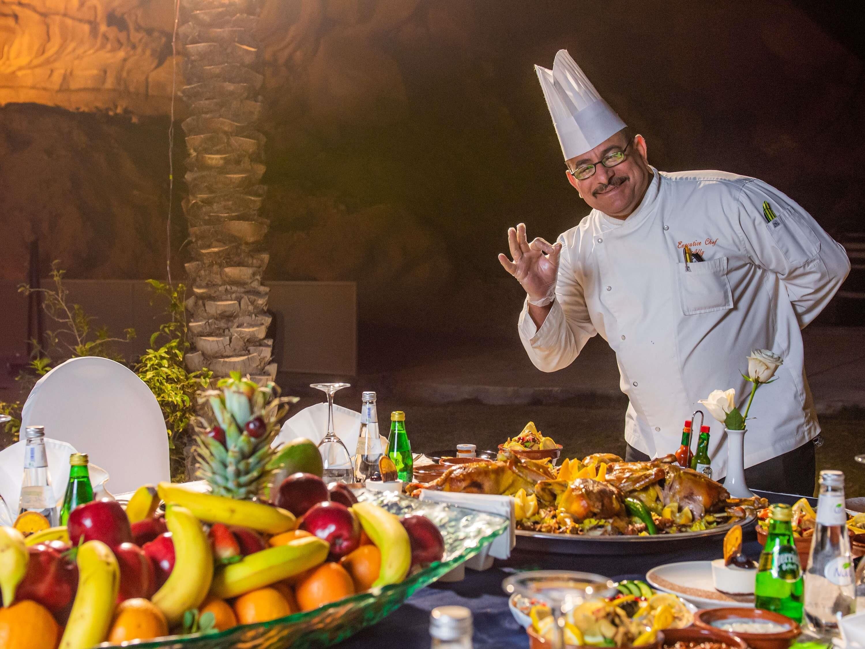 Een kok in een witte uniform met een hoge koksmuts, staand naast een tafel met diverse gerechten en een fruitschaal.