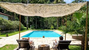 A poolside area with a clear blue pool, two lounge chairs, a woven canopy, and a stone fire pit. - Fazenda Ressaca: Natural Luxury (Ubatuba)