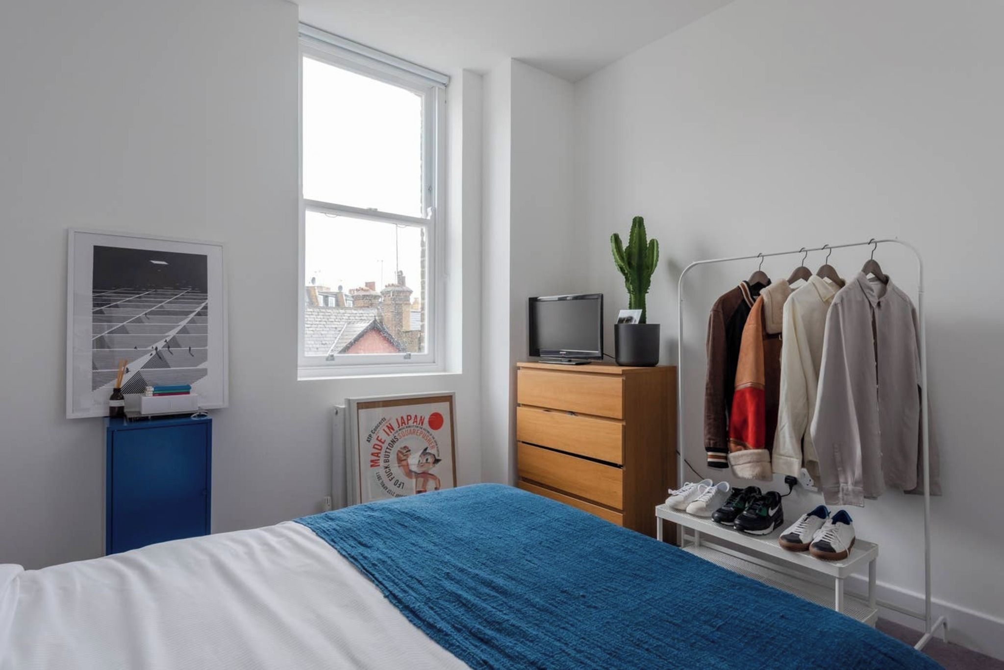 A bedroom with a bed, a dresser, a TV, a cactus, a clothing rack, and a poster on the wall.