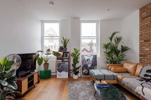 A modern living room with a TV, sofa, and coffee table. There are two windows with city views, a brick wall, and several potted plants. - Hackney Haven Modern 1 BDR Wifi (London)