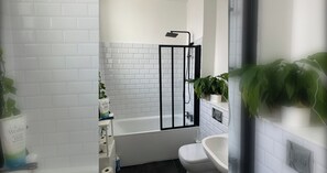 Un baño con cabina de ducha revestida de azulejos blancos, una bañera blanca, un inodoro blanco y plantas en macetas.