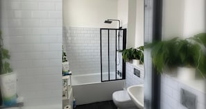 A bathroom with a white-tiled shower enclosure, a white bathtub, and a white sink. There are potted plants on the wall.