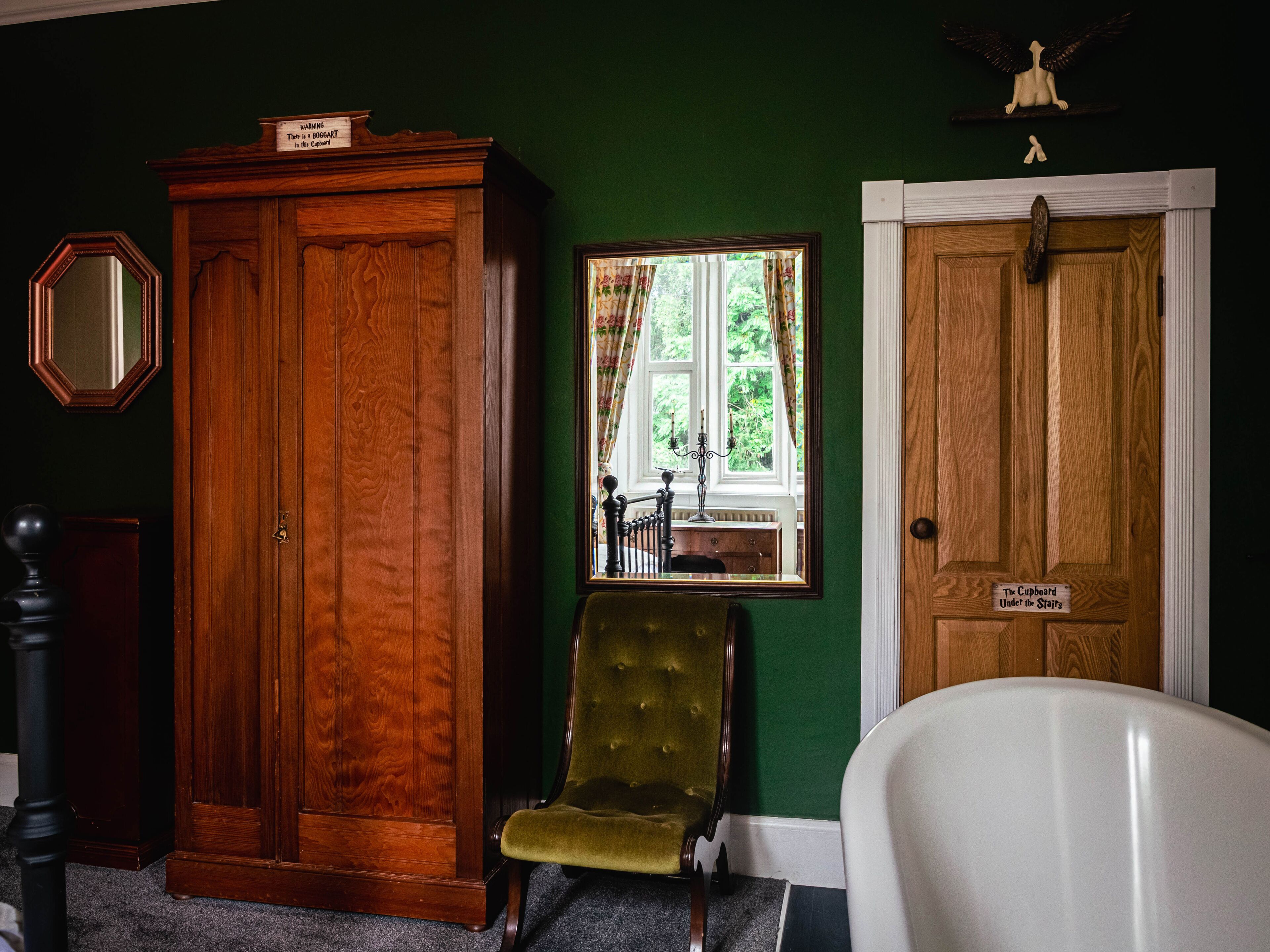 A room with a wooden wardrobe, a green armchair, a white bathtub, and a door labeled "The Cupboard Under the Stairs".