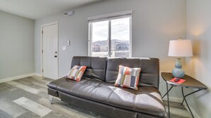 Una sala de estar moderna con un sofá de cuero, dos almohadones decorativos, una mesita de noche con una lámpara y una ventana que ofrece vistas a edificios.