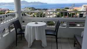 Ein Balkon mit Tisch und Stühlen, mit Blick auf eine Küstenstadt mit Gebäuden und dem Meer.
