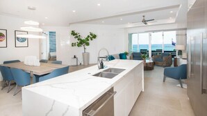 A modern kitchen with a central island, dining area with blue chairs, and a view of the ocean. - Rum Point Club #304 (North Side)