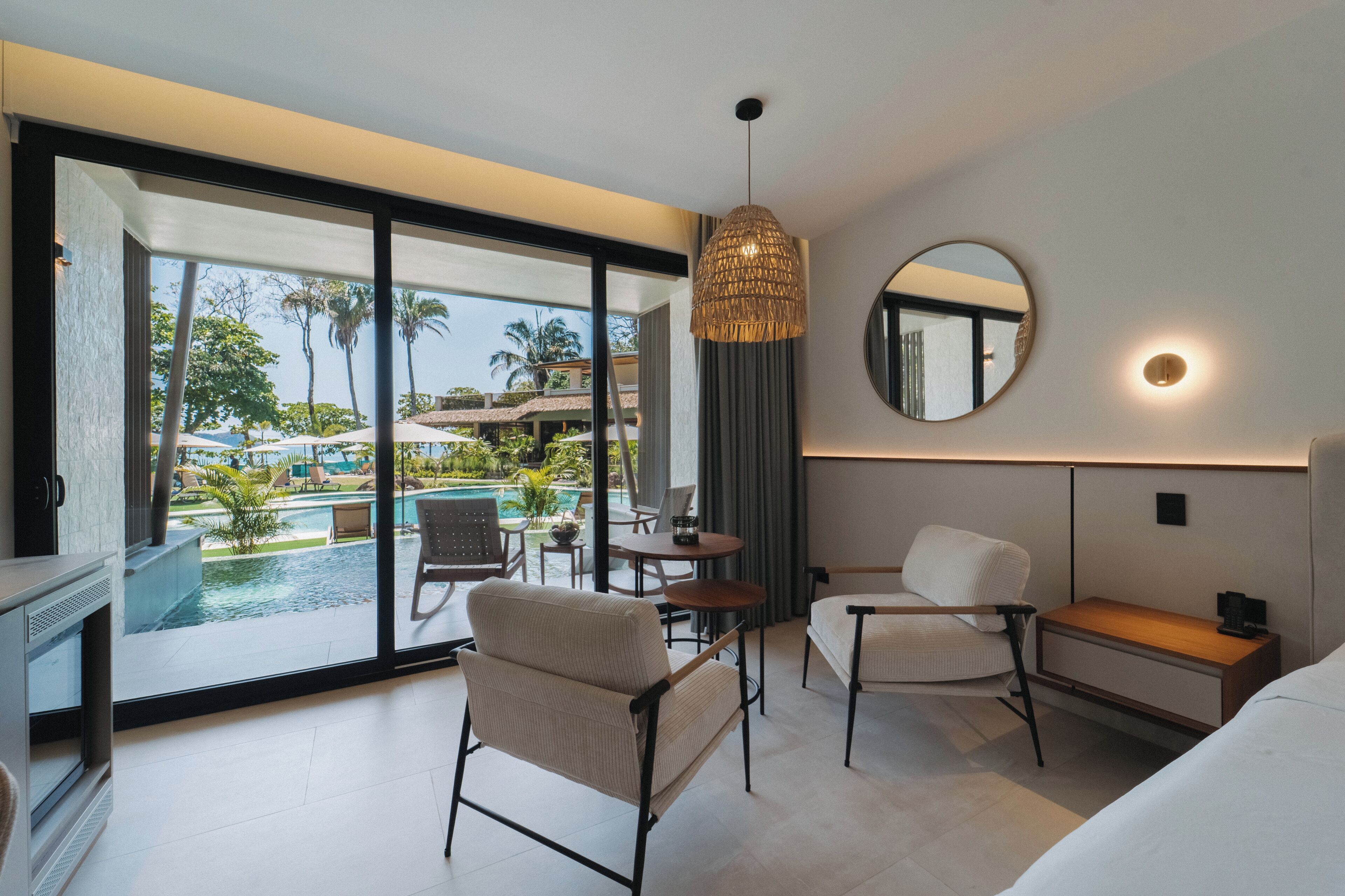 A hotel room with a large sliding glass door to a pool area, a round mirror, a hanging light fixture, and a small table with chairs.
